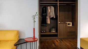 A room with a yellow sofa, a small red table with a plant, a wooden floor, a wardrobe with clothes, and a small table with a radio.
