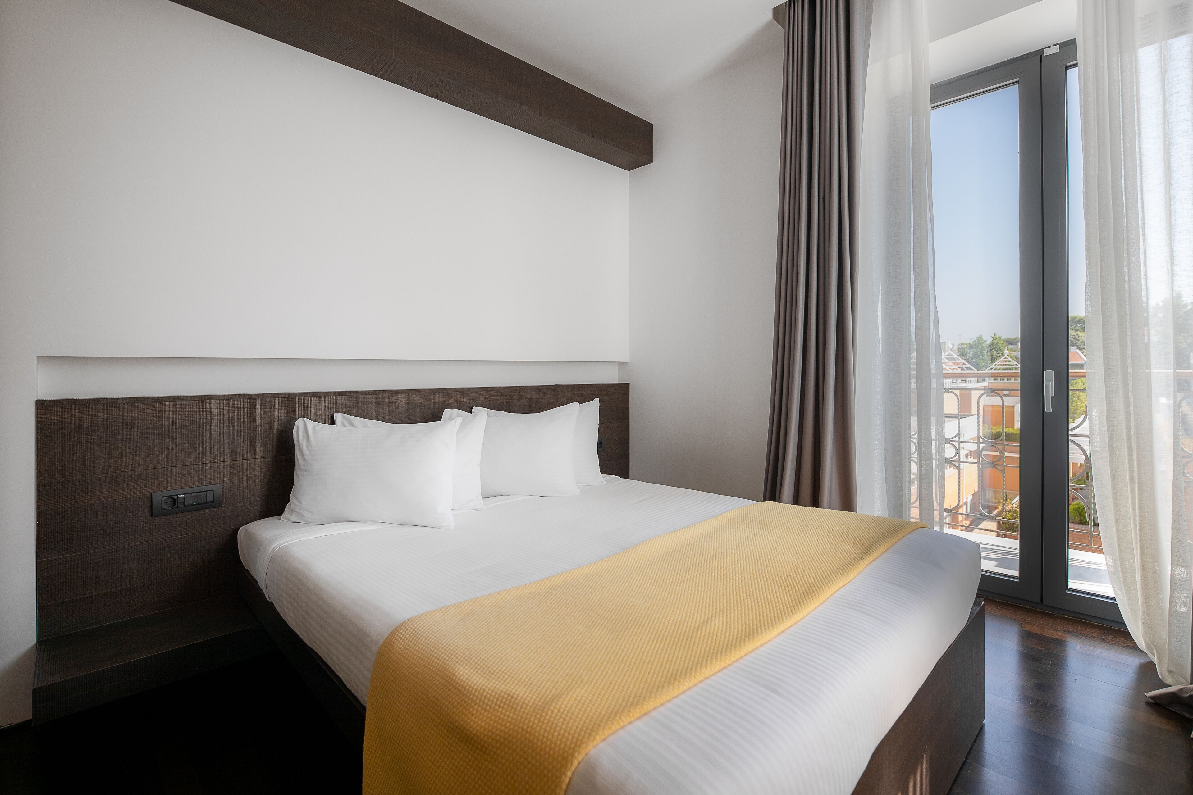 Ein Hotelzimmer mit einem großen Bett, einem Kopfteil, einem Fenster mit Vorhängen und Blick auf Gebäude.
