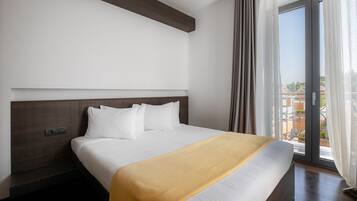 Ein Hotelzimmer mit einem großen Bett, einem Kopfteil, einem Fenster mit Vorhängen und Blick auf Gebäude.