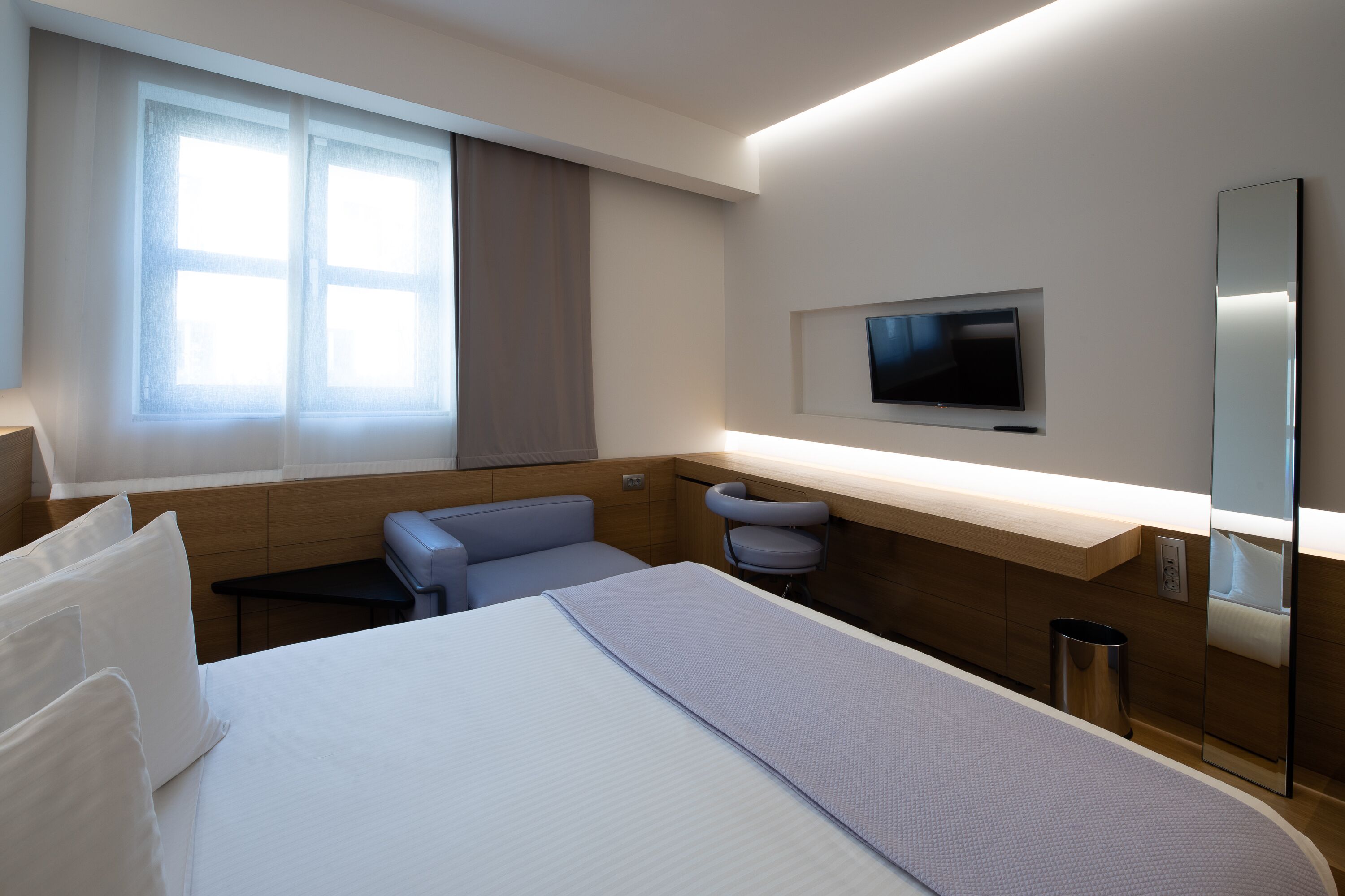 A modern hotel room with a bed, a seating area, a television, and a window with curtains.