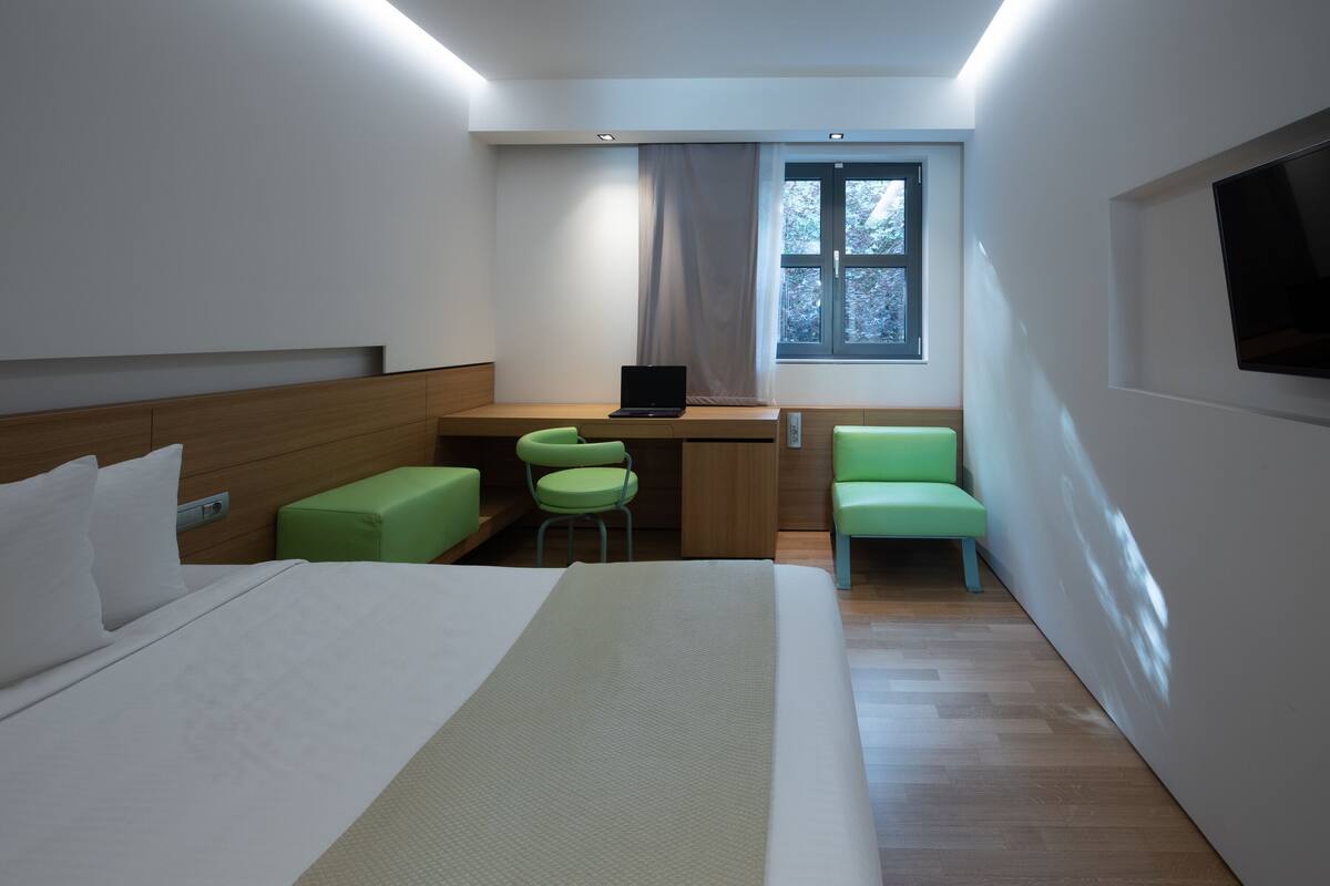 A modern hotel room with a bed, a desk, two green chairs, a laptop, and a TV.