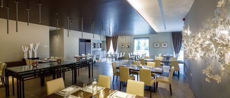 A spacious dining area with multiple tables and chairs, modern lighting fixtures, and a large window providing natural light.