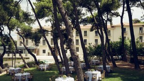Un área de comedor al aire libre con mesas y sillas redondas, rodeada de árboles y un edificio al fondo.