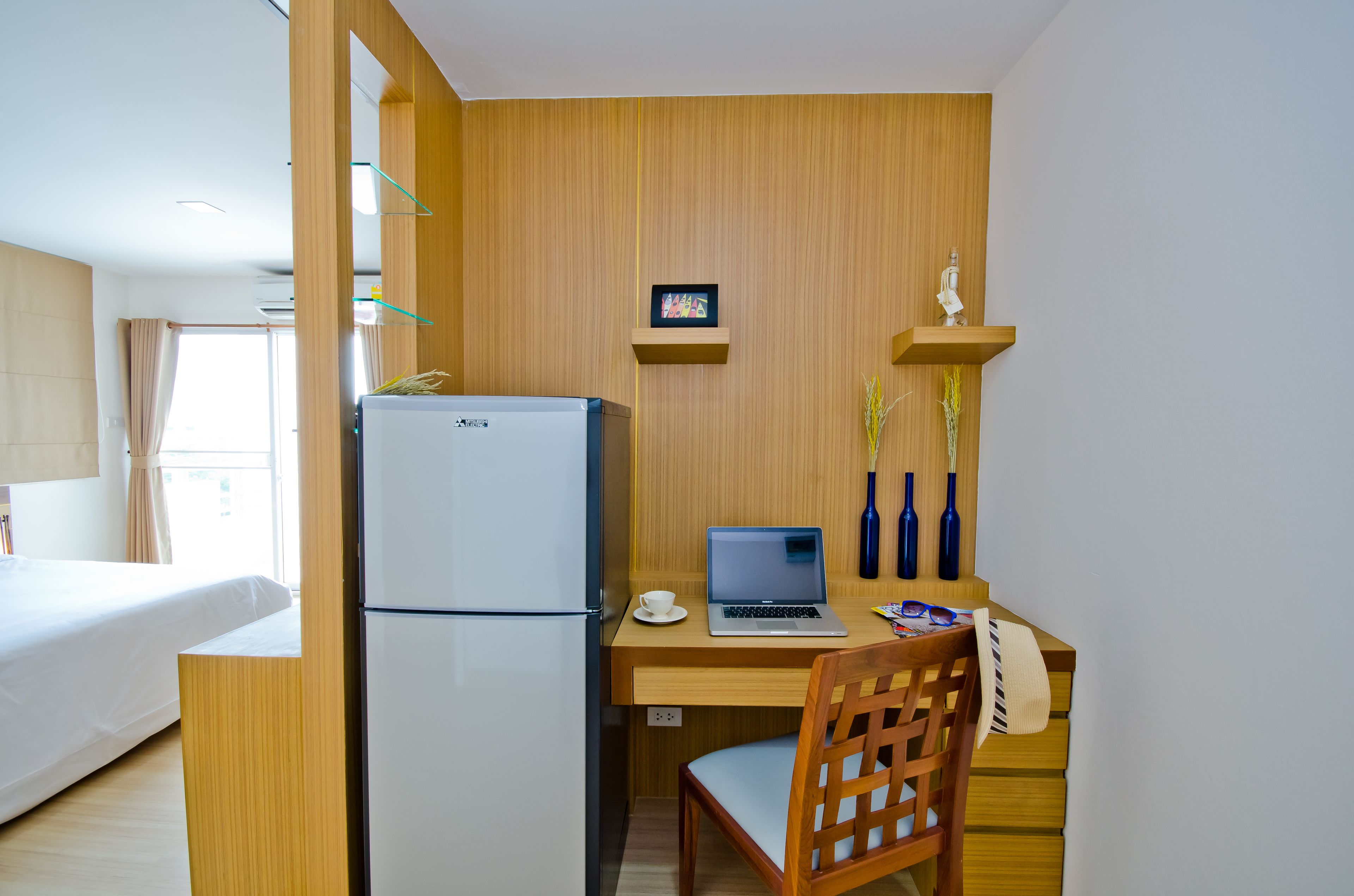A hotel room with a bed, a desk with a laptop, a refrigerator, and a small shelf with decorative items.