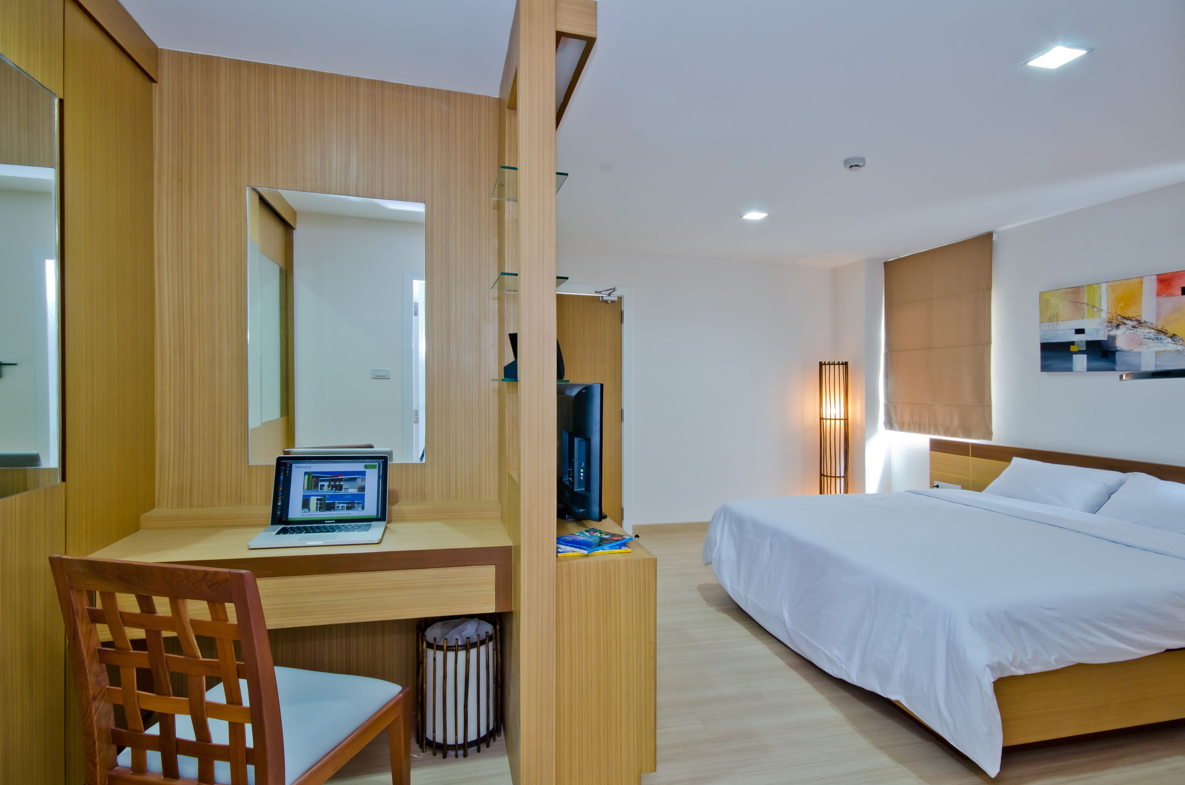 A modern hotel room with a wooden desk, a laptop, a bed with white linens, and a large mirror.