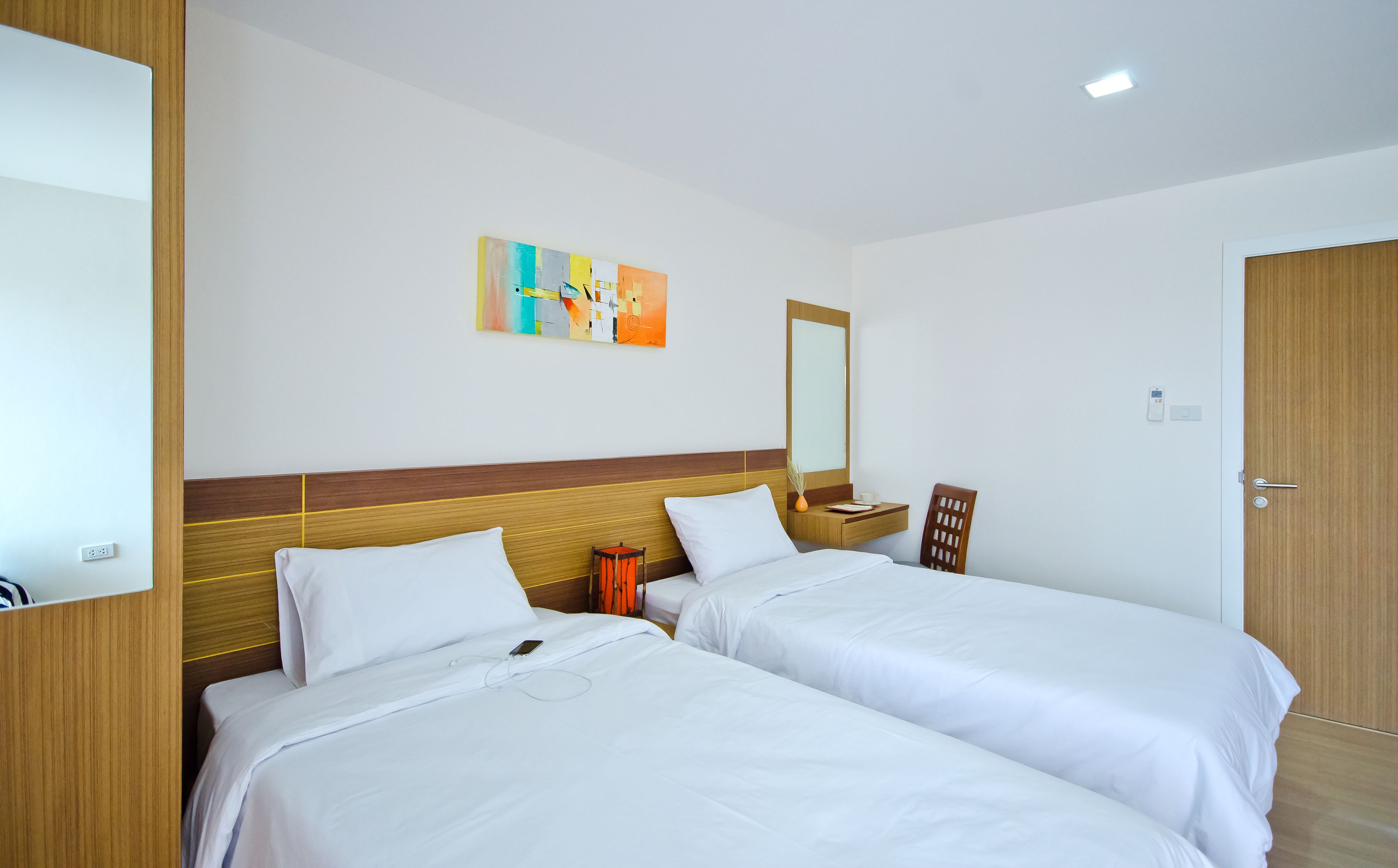 A hotel room with two beds, a wooden headboard, a bedside table with a lamp, a mirror, and a colorful abstract painting on the wall.