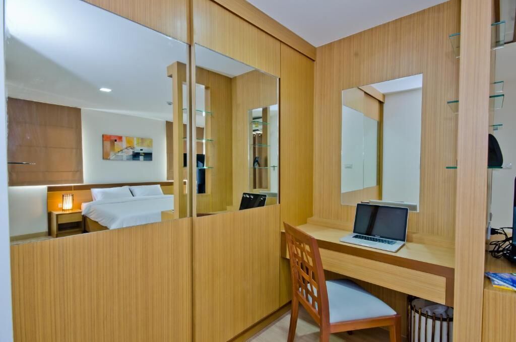 Une chambre d’hôtel avec un bureau en bois, un ordinateur portable, une chaise, un lit, une table de chevet, une lampe et un grand miroir.