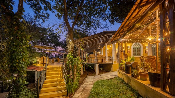 A well-lit outdoor area with a wooden staircase leading to a covered seating area, surrounded by lush greenery and potted plants. - Wildflower Villas (Candolim)