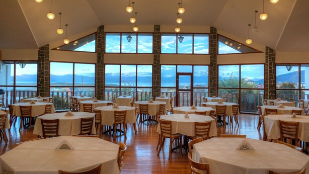 Un restaurante con varias mesas y sillas, amplias ventanas con vistas a las montañas y lámparas colgantes.