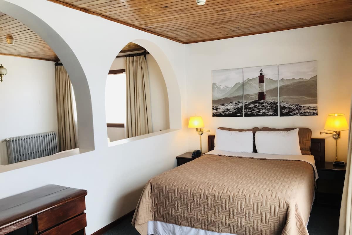 Habitación de hotel con una cama grande, dos lámparas de noche, un escritorio de madera, un radiador y un cuadro de un faro.