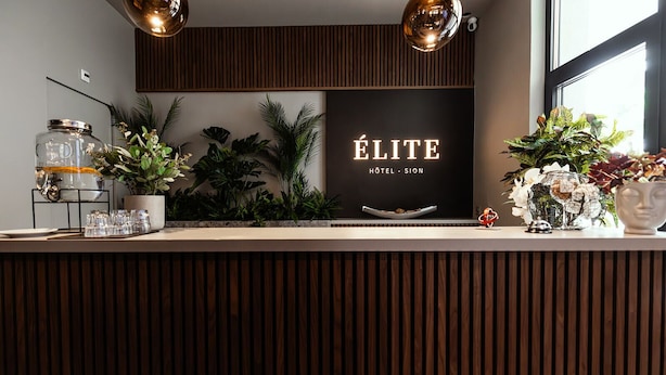 A reception desk with a sign reading 'ÉLITE HOTEL - SION,' surrounded by plants and decorative items.
