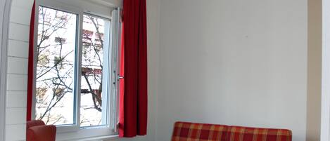 Una habitación con un sofá rojo a cuadros, un sillón de madera mecedora, un pequeño escritorio de madera y una ventana con cortinas rojas.