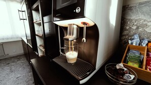 A modern espresso machine with a glass cup underneath, a coffee grinder, and a shelf with coffee pods and a basket of beer bottles. - Hôtel Elite (Sion)