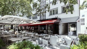 An outdoor seating area with tables and chairs, a red awning, and a building with 'HOTEL ÉLITE' signage.