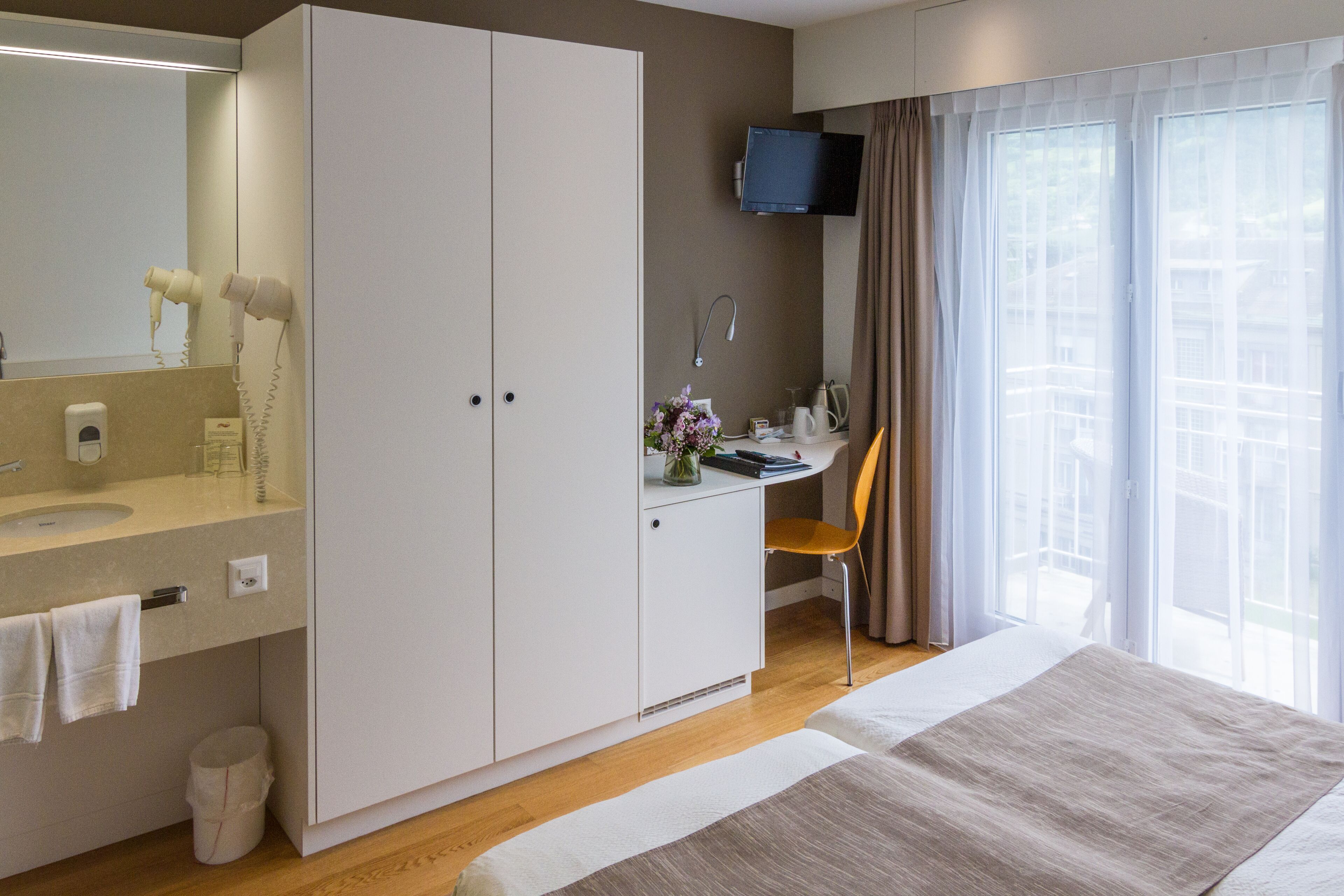Ein Hotelzimmer mit großem Kleiderschrank, Schreibtisch mit Stuhl, Wandfernseher und einem großen Fenster mit Vorhängen.
