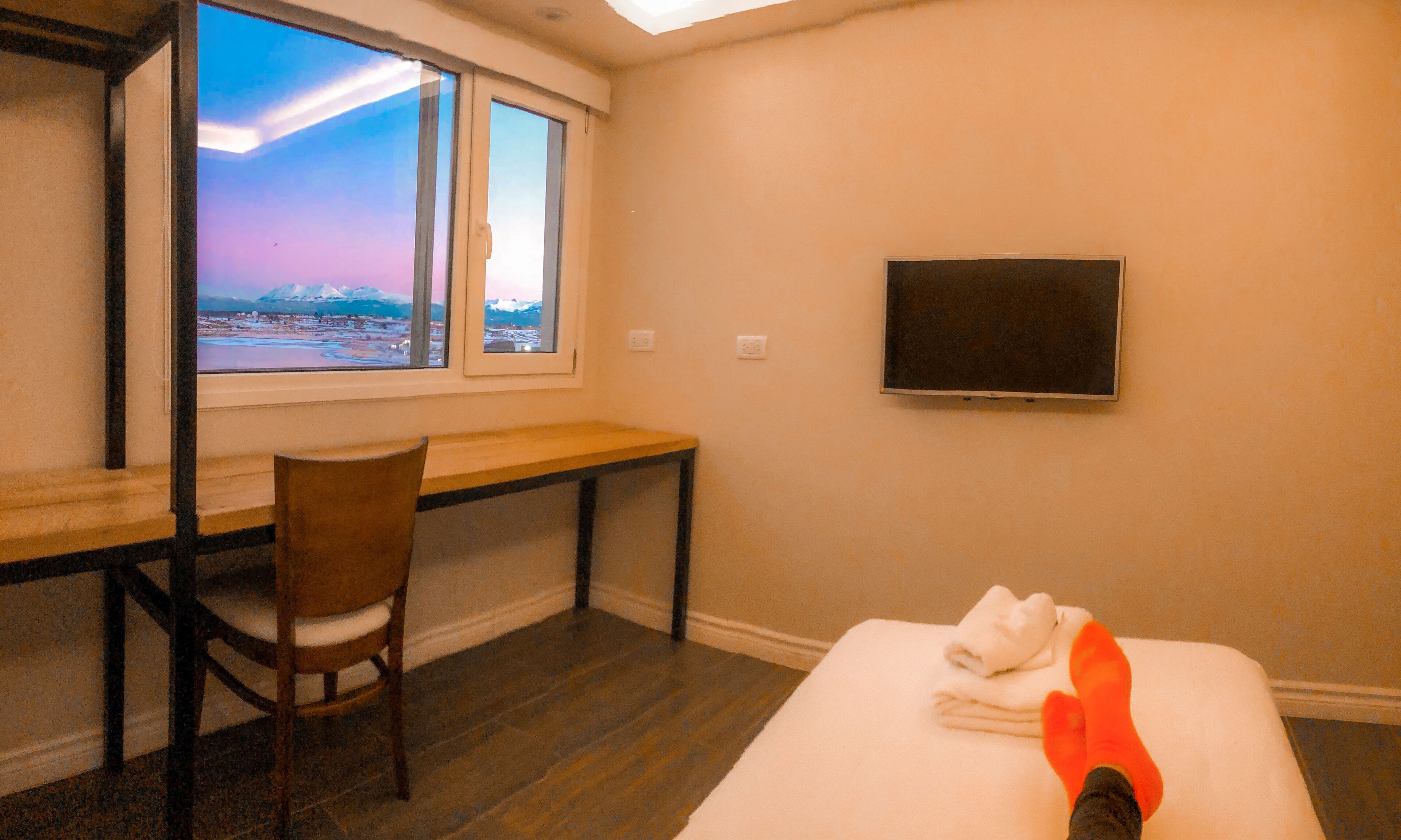 Une chambre d’hôtel avec un bureau en bois, une chaise, un lit avec une serviette blanche et un téléviseur fixé au mur.