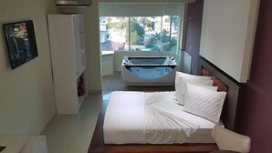 A hotel room with a bed, a chair, a small table, a bathtub, and a window with a view of buildings. - Emirates Hotel & Suítes (Santana do Livramento)