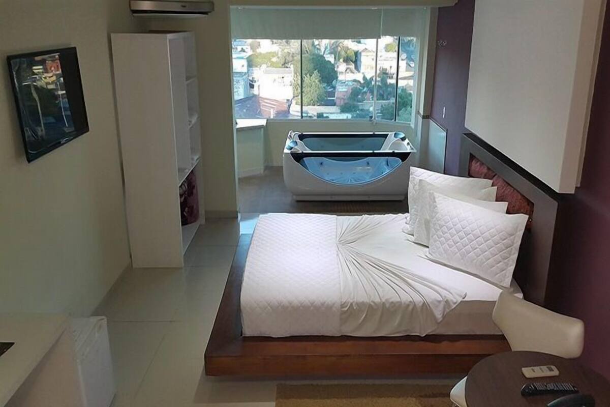 Een hotelkamer met een bed, een stoel, een tafeltje, een badkuip en een raam met uitzicht op gebouwen.