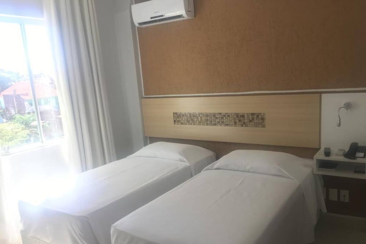 Een hotelkamer met twee enkele bedden, een houten hoofdbord, een klein bureau met telefoon en een raam met gordijnen.