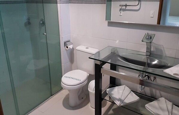 Un baño con cabina de ducha de vidrio, inodoro blanco y lavamanos de vidrio con un espejo arriba.