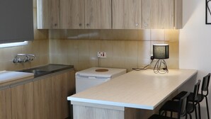 A modern kitchen with wooden cabinets, a white countertop, a built-in dishwasher, and a bar area with stools. - Layiotis Hotel Apartments (Larnaca)