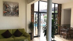 A spacious living area with a green sofa, a glass coffee table, and a dining area with wooden chairs and a table. There is a large window with a view of the outside. - New York Plaza Hotel Apartments (Paphos)