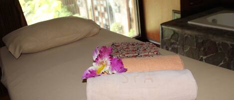 A neatly made bed with a floral-patterned towel and a rolled towel, a window with a view of greenery, and a stone bathtub.