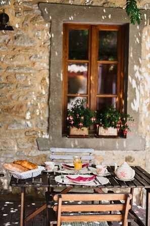 Una rústica pared de piedra con un ventanal de madera, una mesa dispuesta con pan, platos y una tetera, y plantas en macetas en el alféizar.