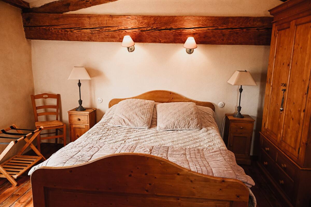 A wooden bed with a quilt, two bedside lamps, a wooden chair, a small wooden nightstand, and a wooden wardrobe.