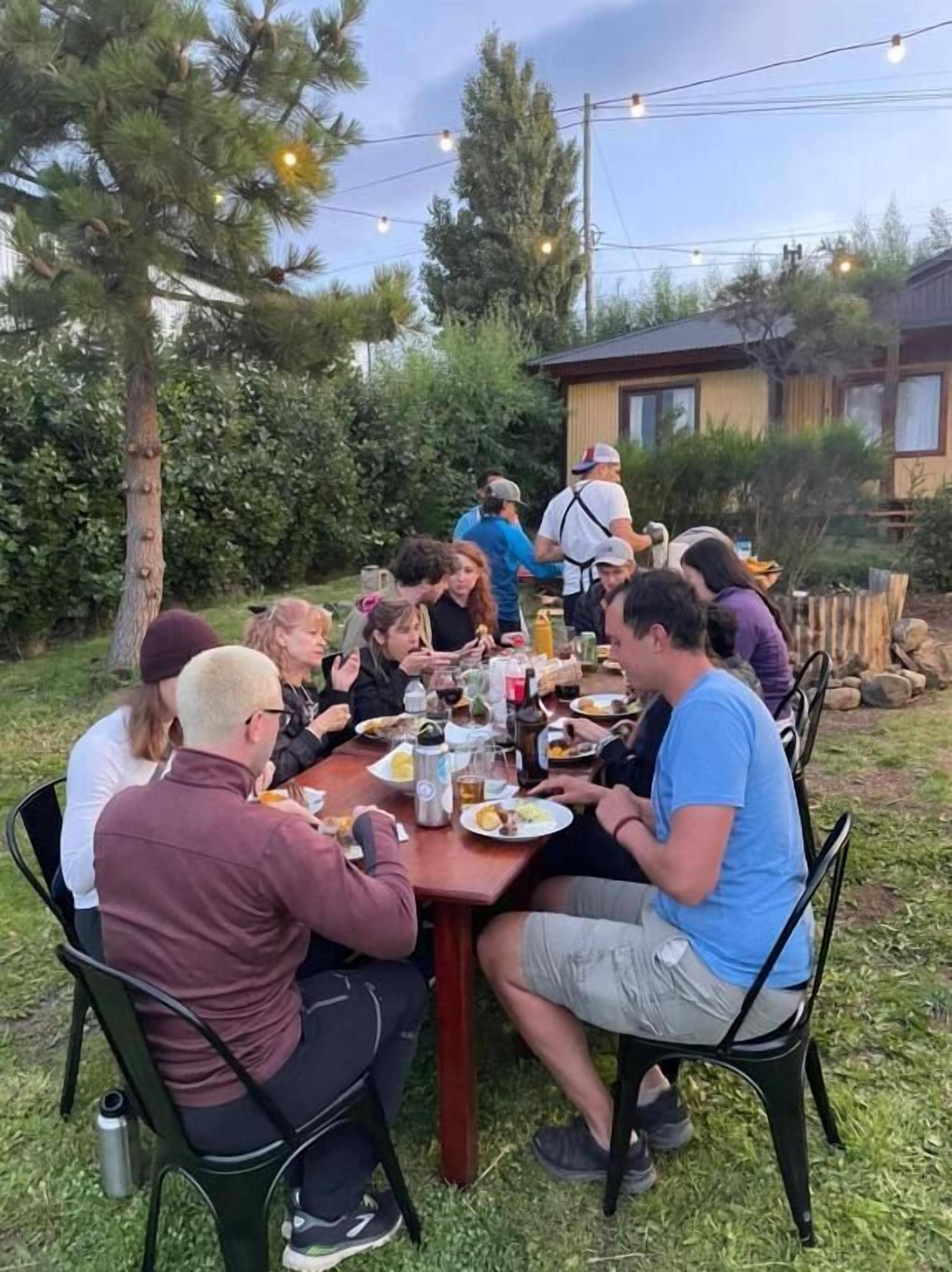 A group of people dining outdoors at a wooden table with chairs, surrounded by greenery and a house in the background.