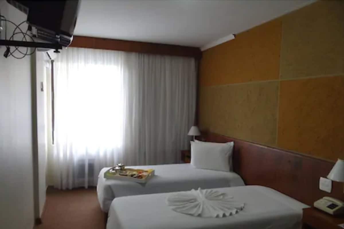 Une chambre d’hôtel avec deux lits, une télévision, une fenêtre avec des rideaux et une table de chevet avec une lampe.