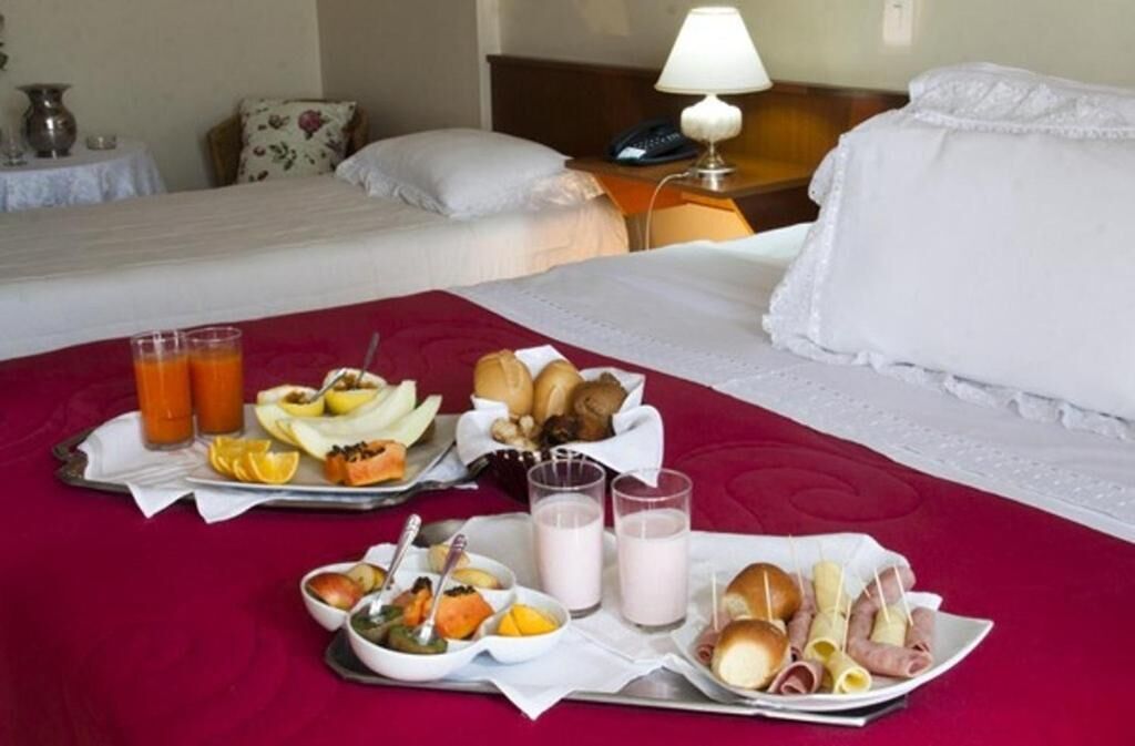 Habitación de hotel con dos camas, un carrito de desayuno con frutas, pasteles y bebidas, y una mesita de noche con una lámpara.