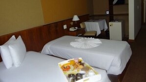 Quarto de hotel com duas camas, uma mesa pequena com abajur, um refrigerador e um porta-roupas com peças penduradas.