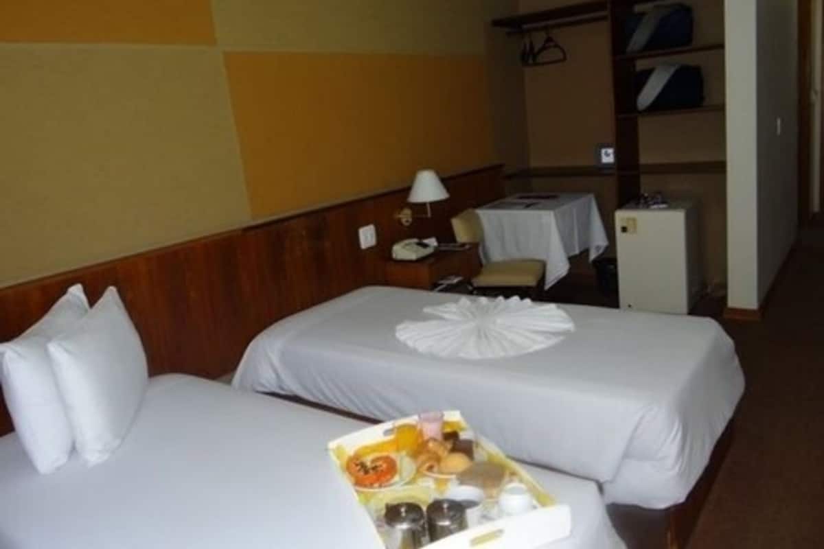 Une chambre d’hôtel avec deux lits, une petite table avec une lampe, un réfrigérateur et un porte-manteau avec des vêtements suspendus.