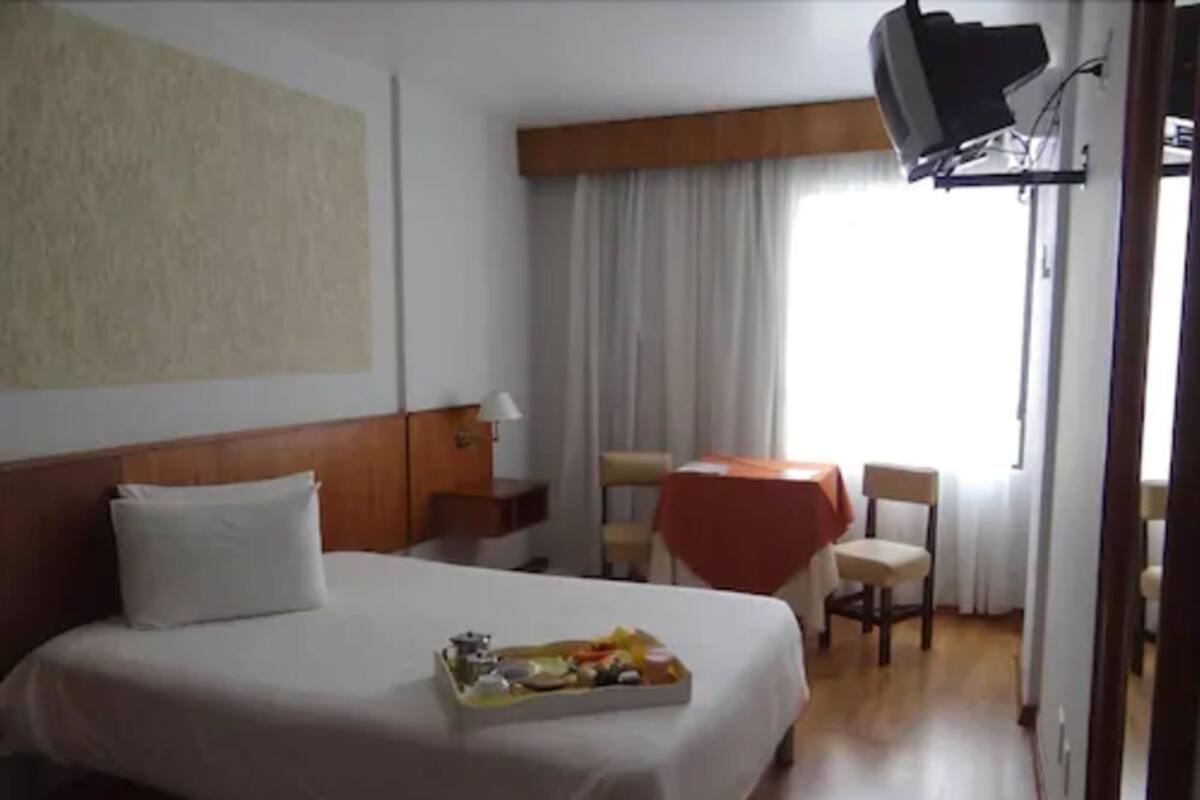 Une chambre d’hôtel avec un lit, une petite table recouverte d’une nappe rouge, un téléviseur fixé au mur et une fenêtre avec des rideaux.