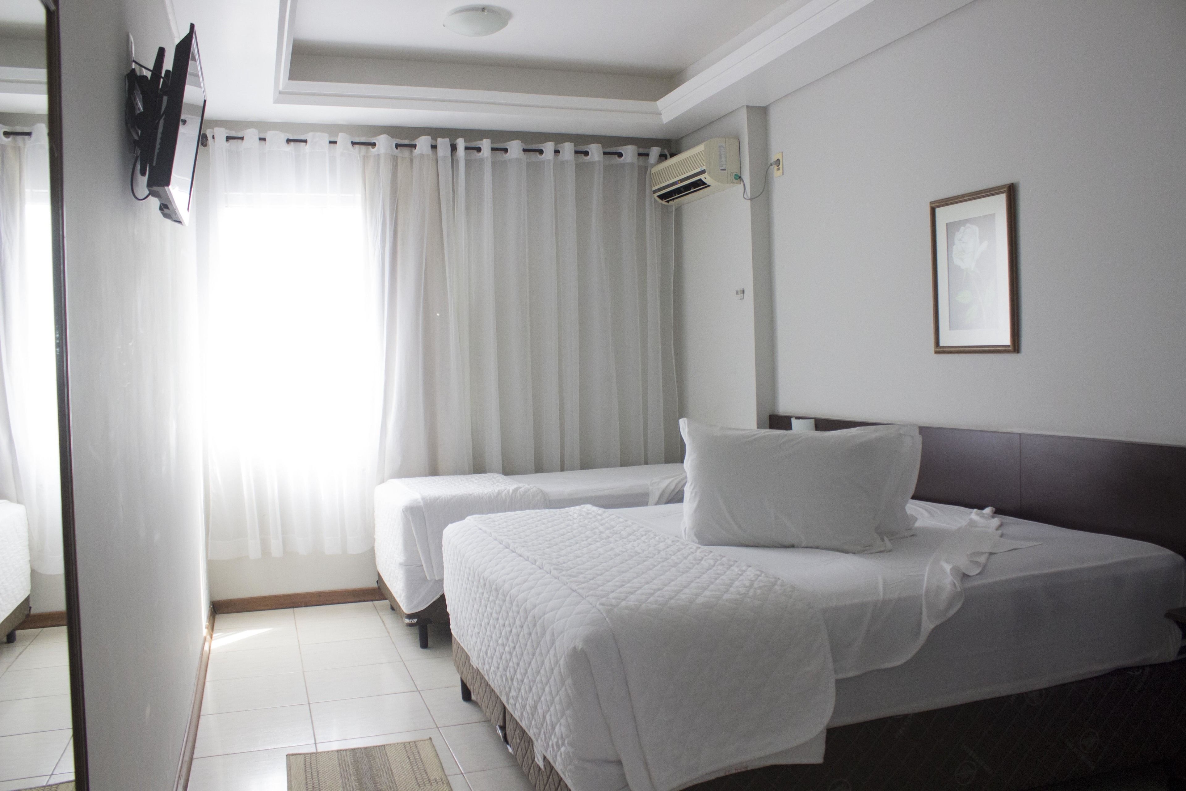 A hotel room with two beds, a large window with sheer curtains, a wall-mounted air conditioner, and a framed picture on the wall.
