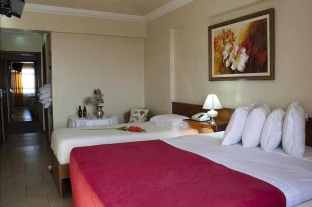 Een hotelkamer met twee bedden, een nachtkastje met lamp, een schilderij aan de muur en een gang die naar een andere kamer leidt.