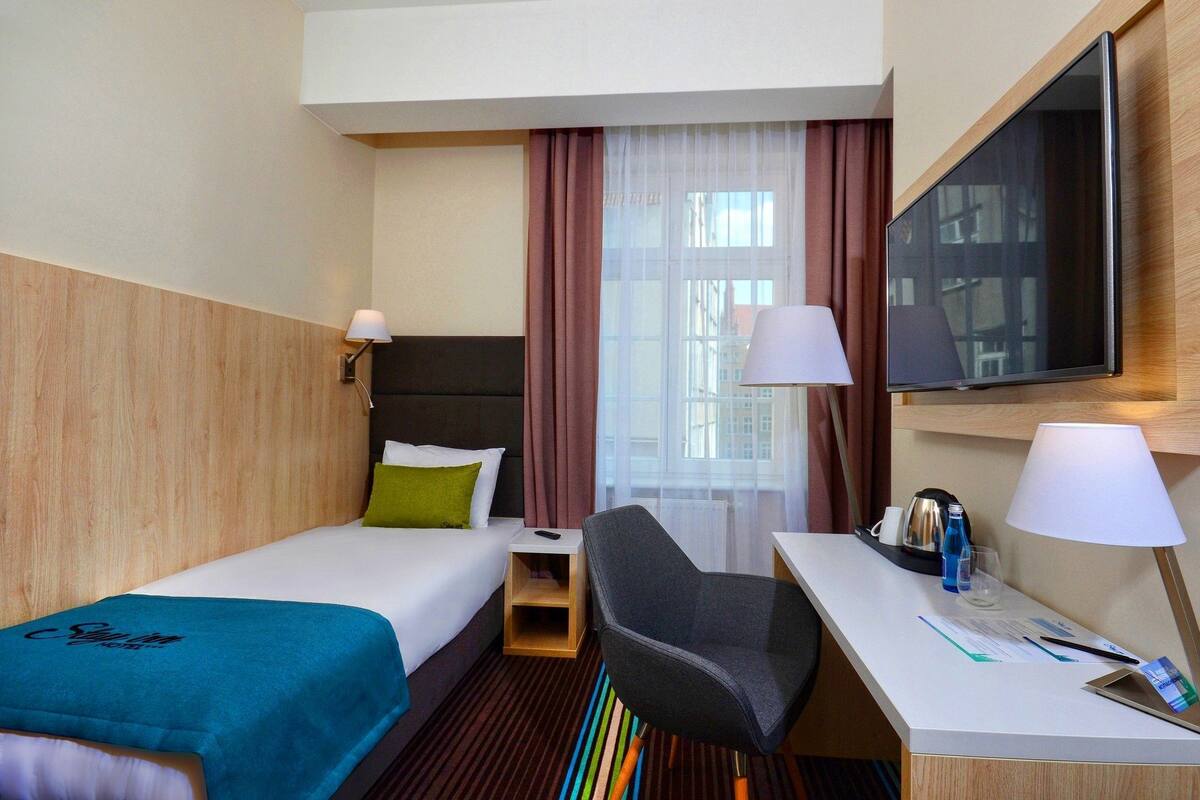 A hotel room with a bed, a desk, a chair, a TV, and a window with curtains.