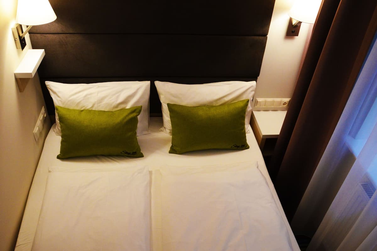 A hotel room with a bed, two green pillows, white bedding, and two bedside lamps.