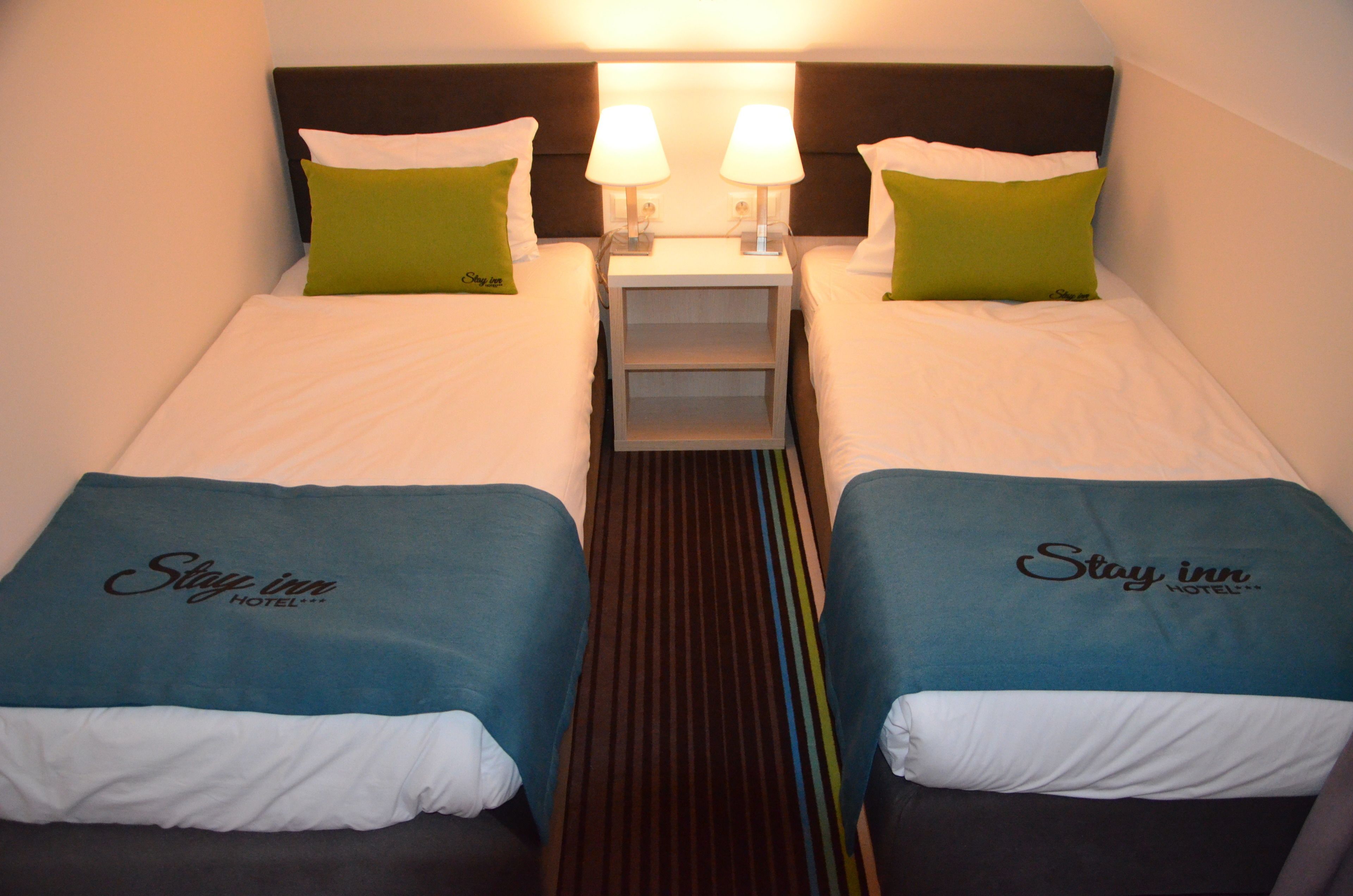 A hotel room with two beds, each with a 'Stay inn' branded bedspread, a nightstand with two lamps, and green pillows.