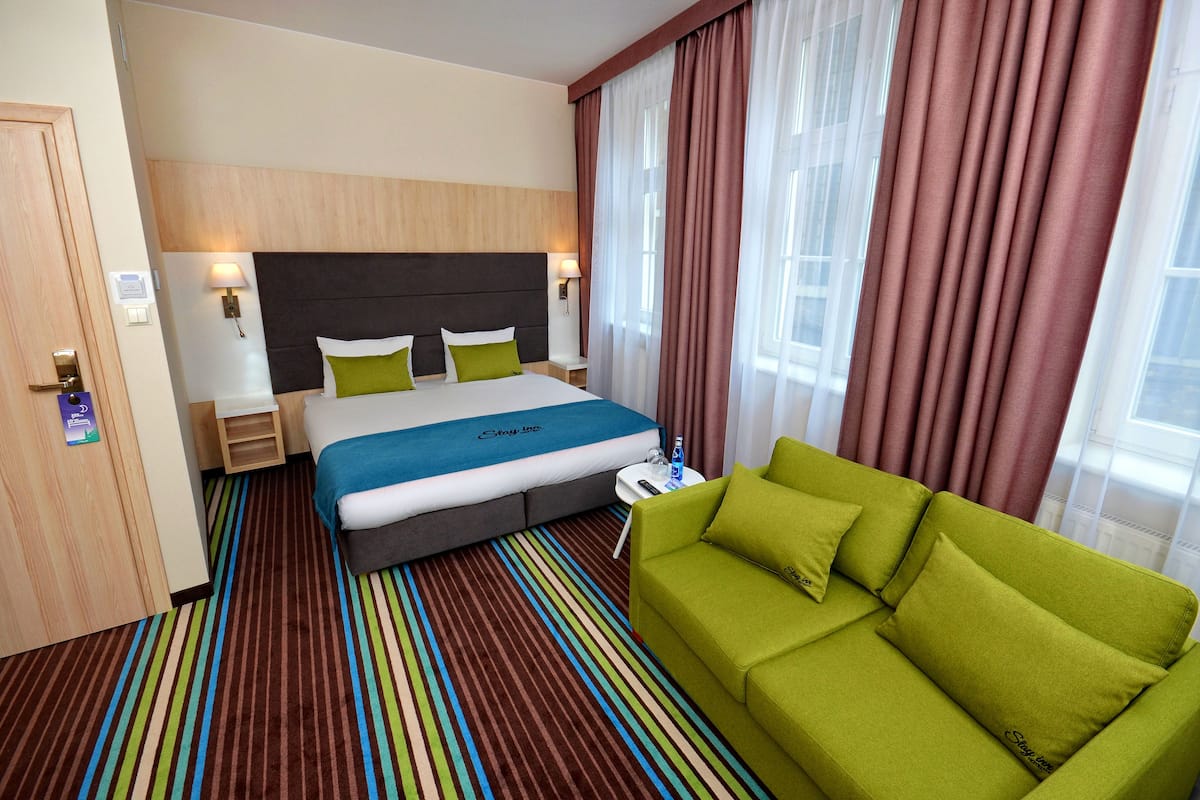 A hotel room with a bed, a green sofa, striped carpet, and a window with curtains.