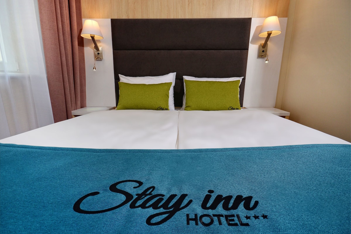A hotel room with a large bed, two pillows, a headboard, and two wall-mounted lamps.