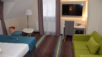 A hotel room with a bed, a green sofa, a desk with a lamp, a TV, and a window with curtains.