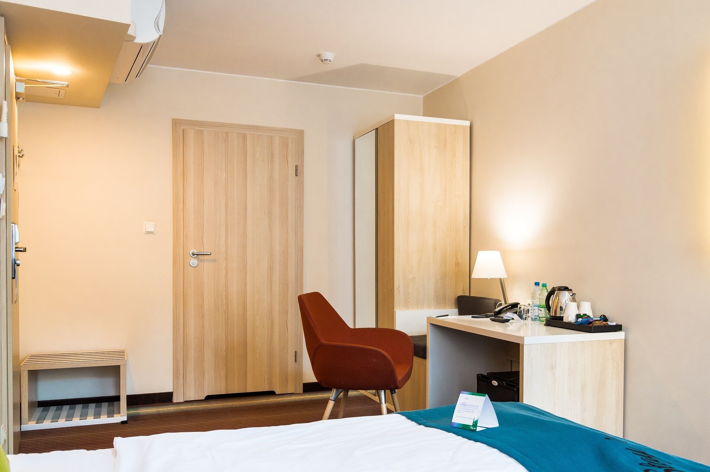 A hotel room with a bed, a desk, a chair, a small table, a mirror, and a lamp.