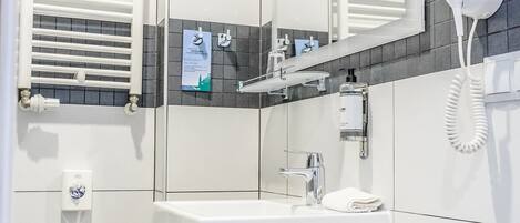 A modern bathroom with a large mirror, a white sink, a towel rack, a hairdryer, and a small trash bin.