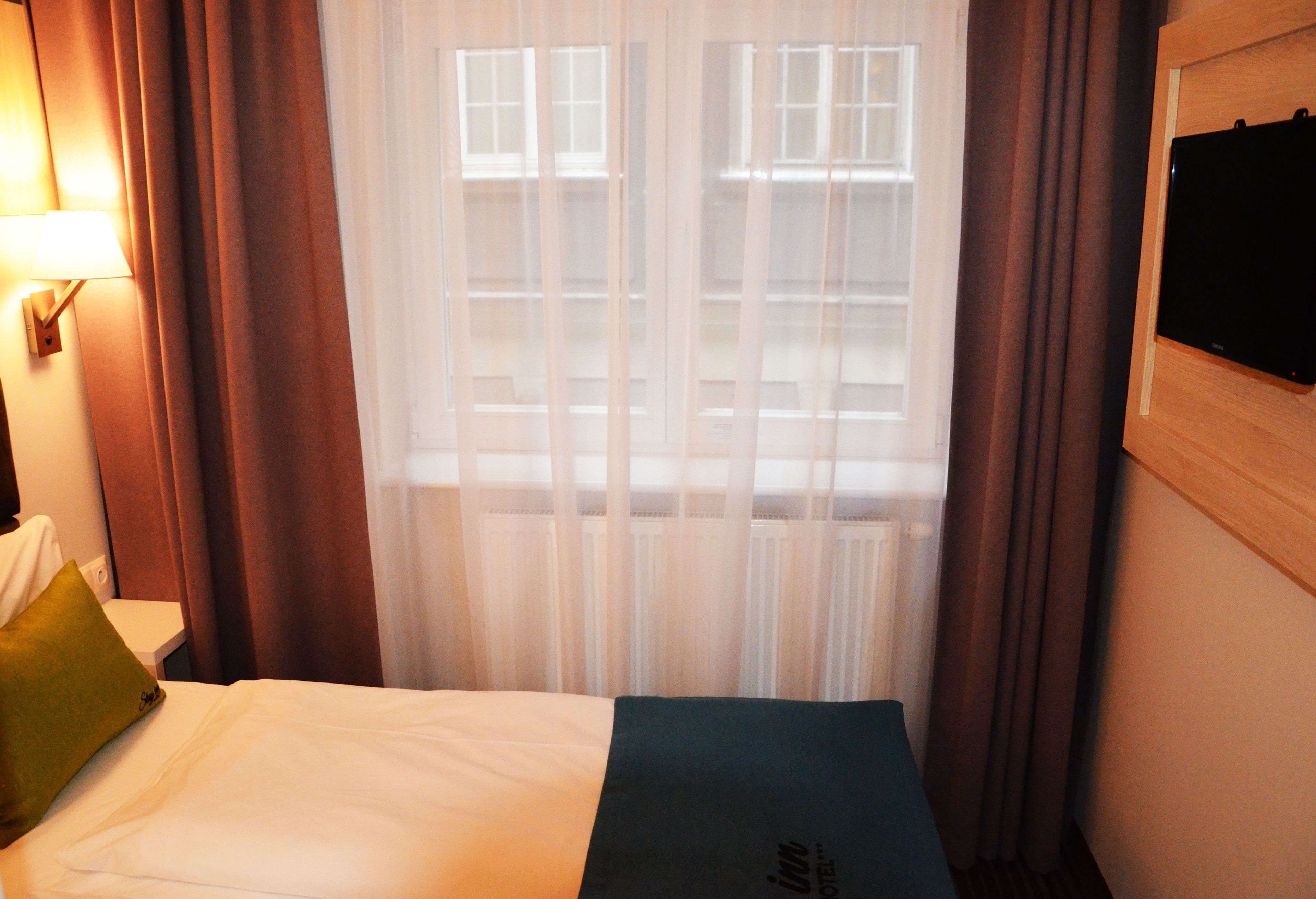 Habitació d'hotel amb un llit, una tauleta de nit, una làmpada i una finestra amb cortines.