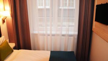Habitació d'hotel amb un llit, una tauleta de nit, una làmpada i una finestra amb cortines.