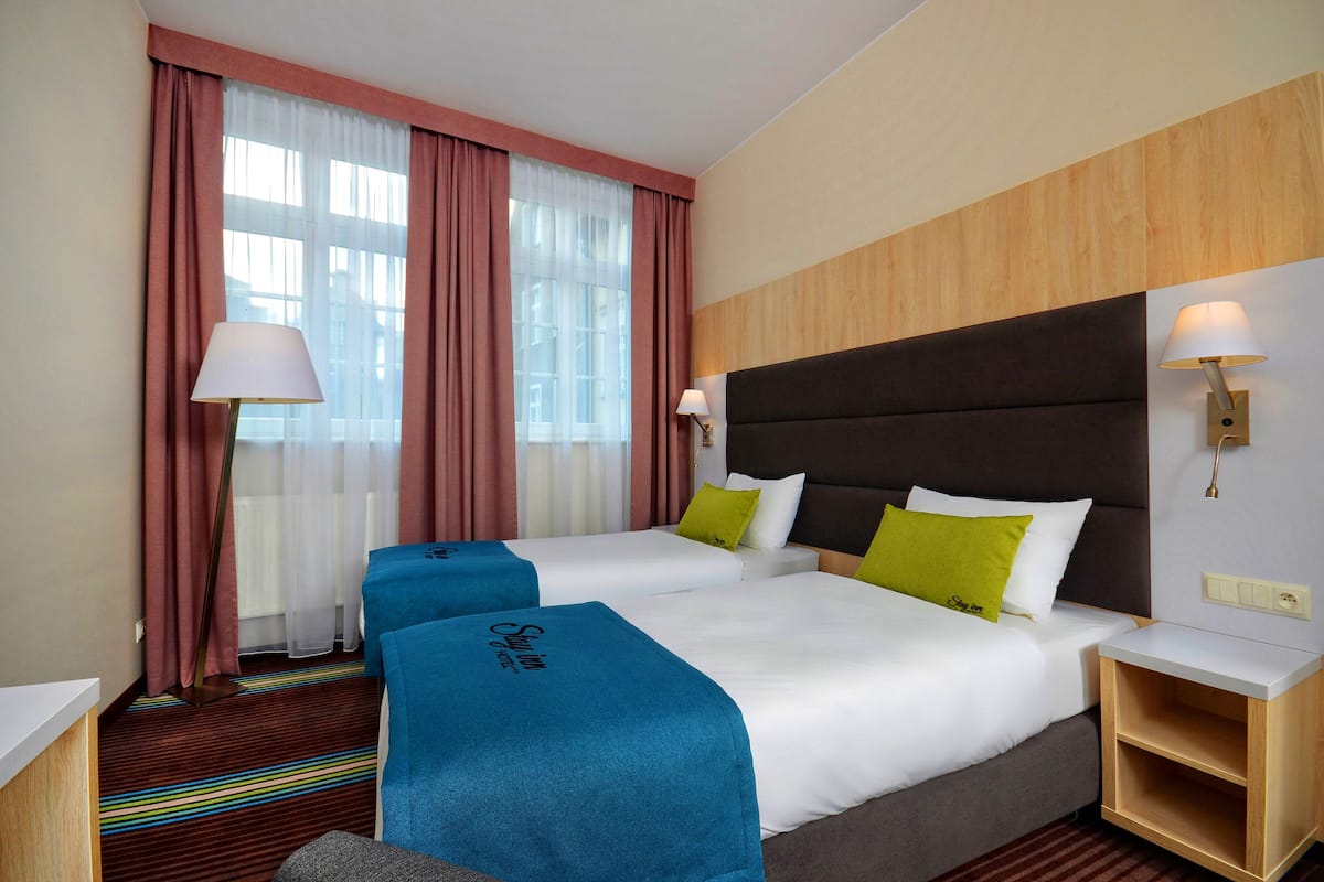 A hotel room with two beds, a large window with curtains, a bedside table with a lamp, and a wooden headboard.