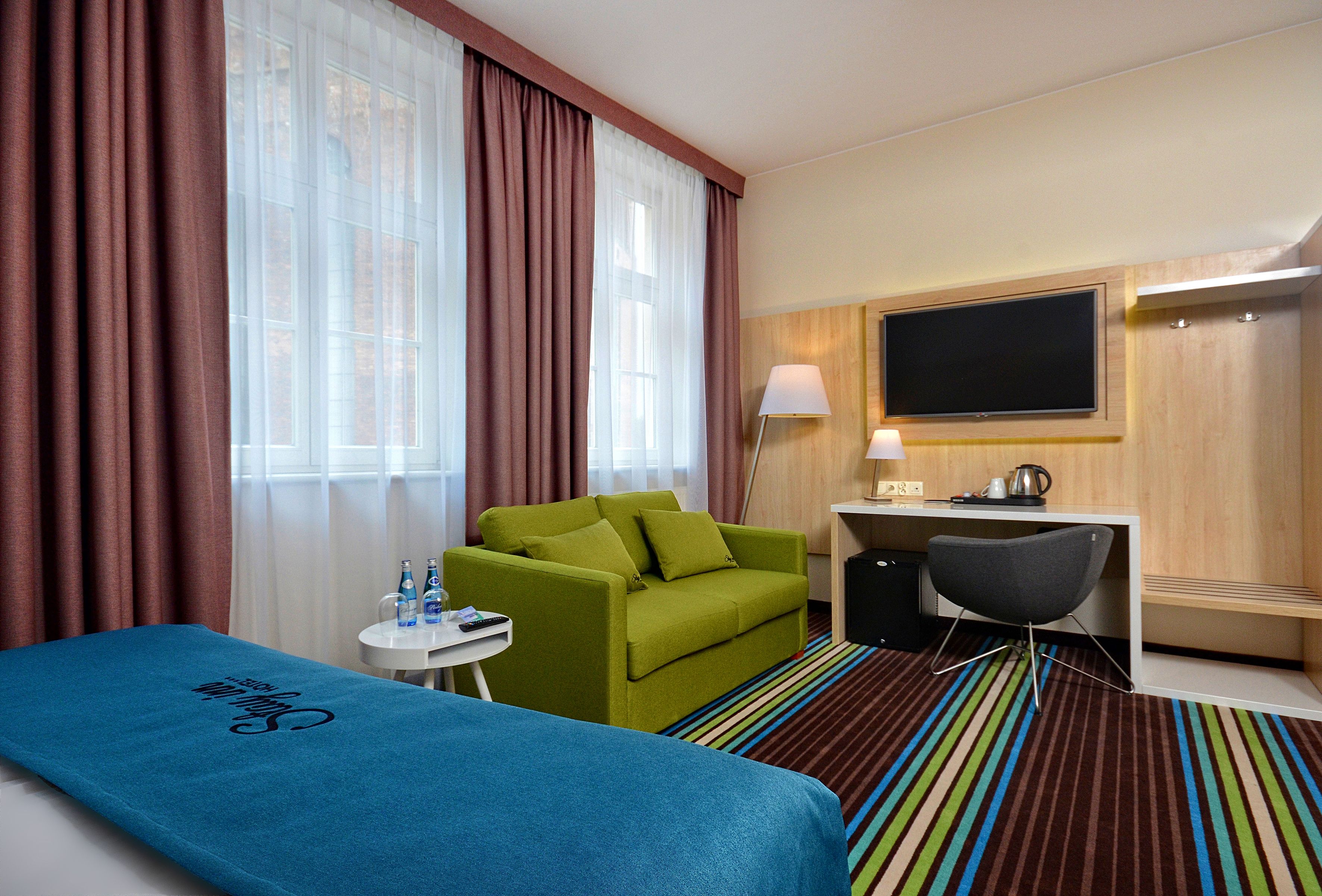 Habitación de hotel con cama, sofá verde, escritorio con silla, televisor y ventana con cortinas.
