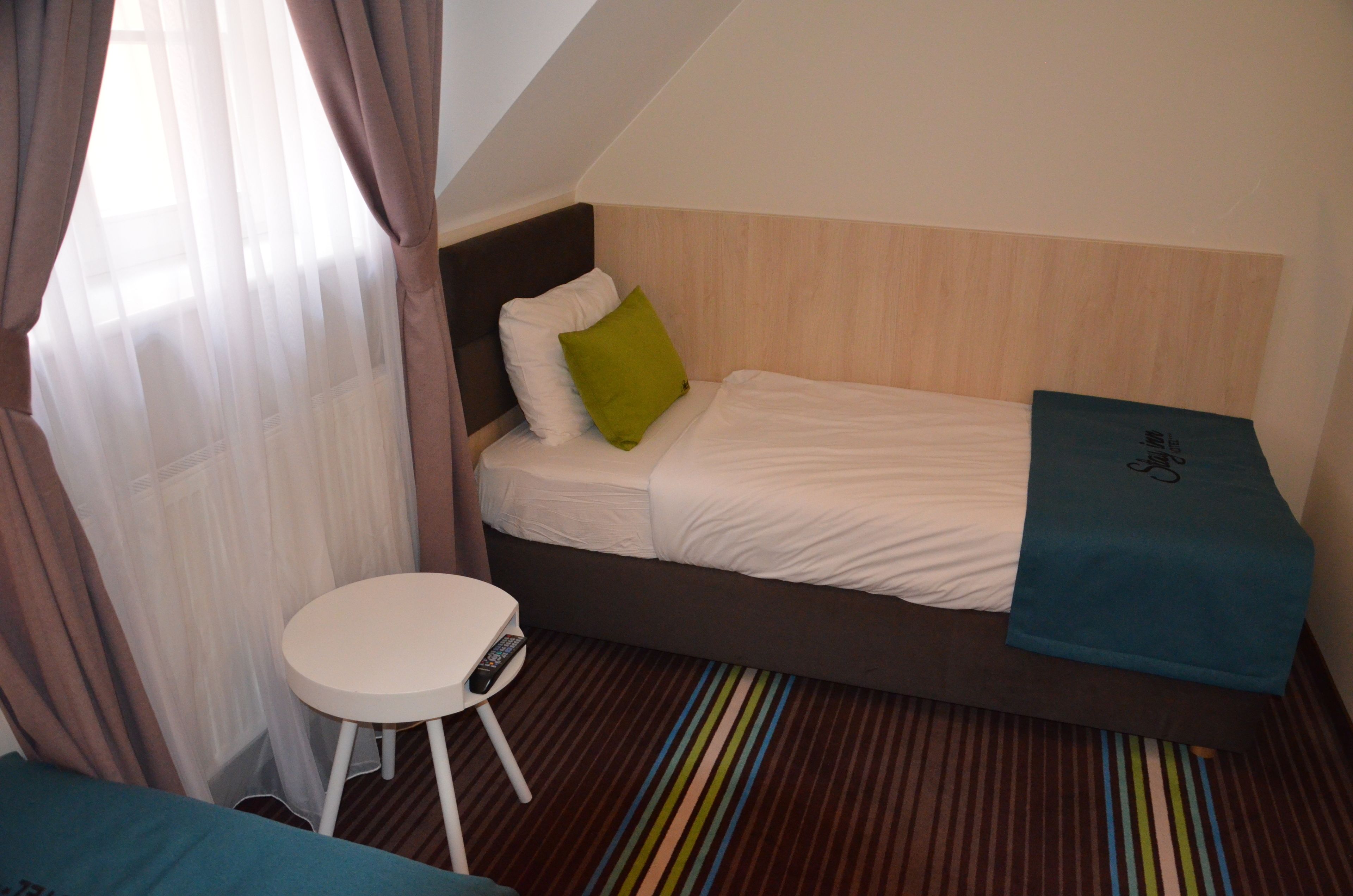 A small, single-bed room with a wooden headboard, a small white table with a remote control, and a colorful striped rug.